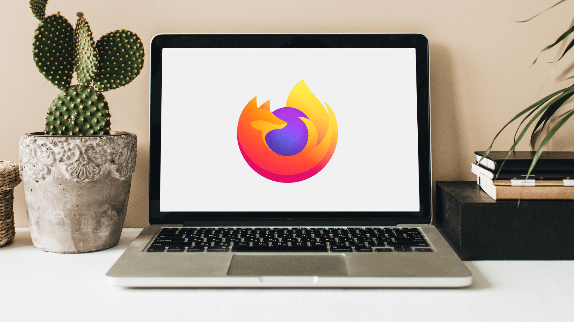Laptop on desk displaying Firefox logo, beside a potted cactus and stack of books, relating to the best browser for Windows 11.